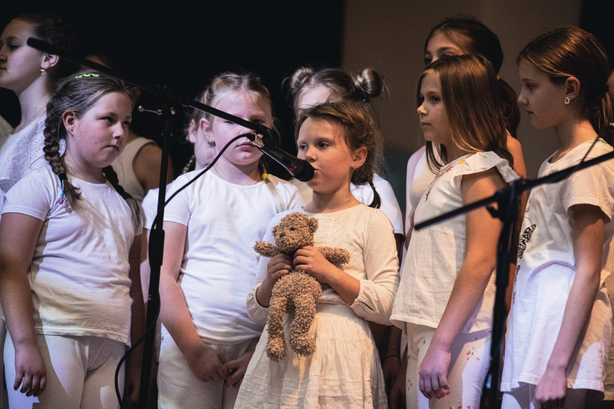 Dziewczynka w białej sukience trzyma kurczowo pluszowego misia śpiewając do mikrofonu. Dookoła niej znajduje się reszta dzieci.